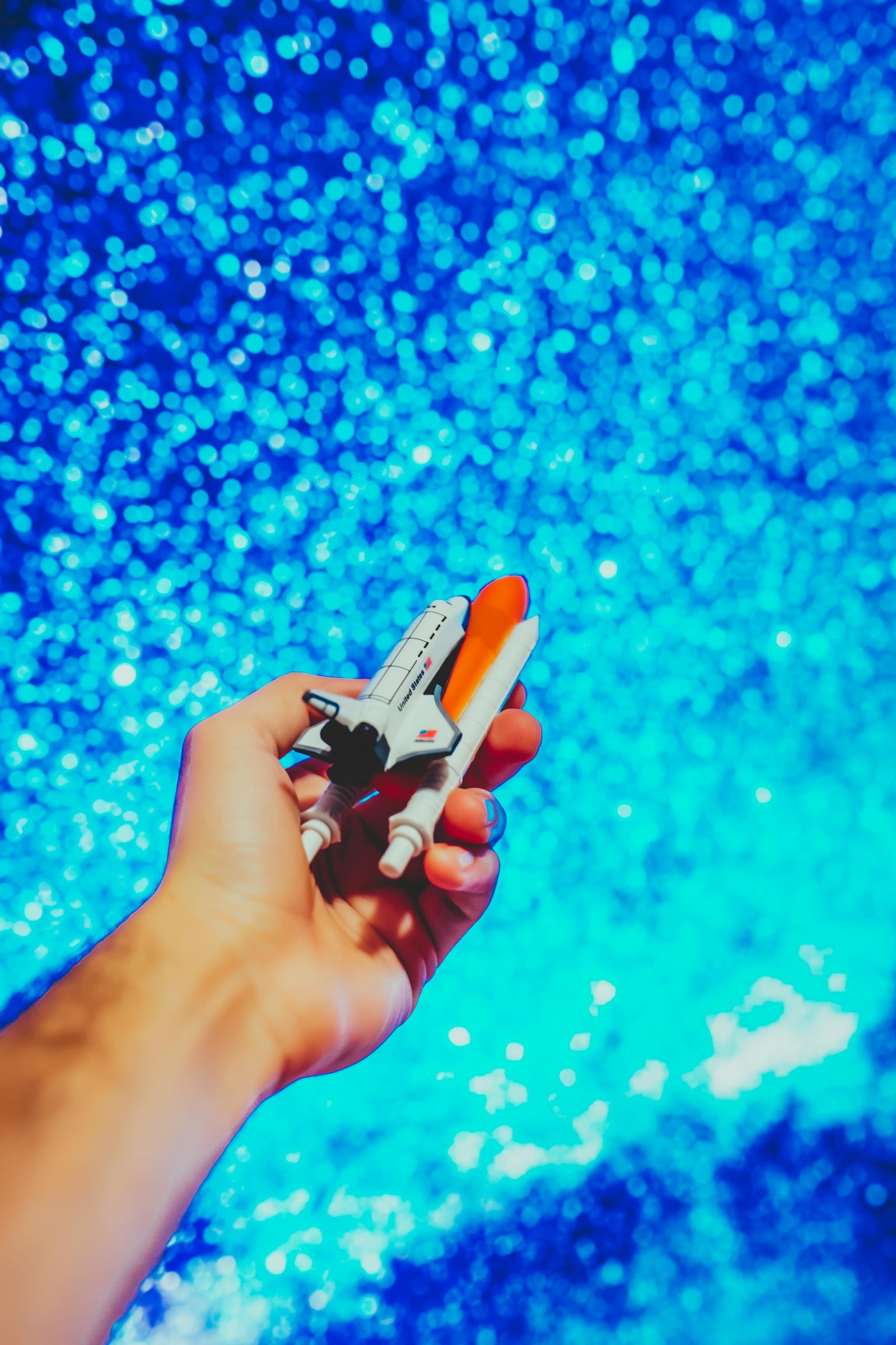 A close-up of a hand holding a toy rocket against a vibrant blue sparkling background.